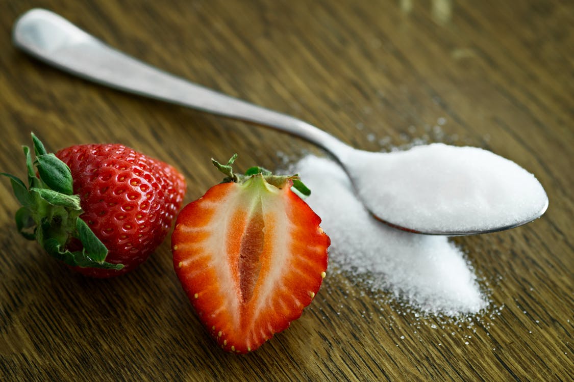 Whole and halved strawberries beside a spoonful of white sweetener on a wooden surface, illustrating the question: are sweeteners bad for you?
