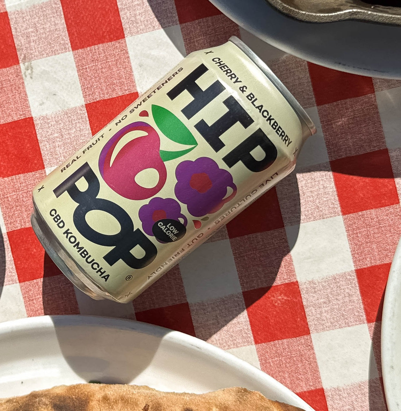A can of HIP POP cherry and blackberry CBD kombucha resting on a red and white checked tablecloth