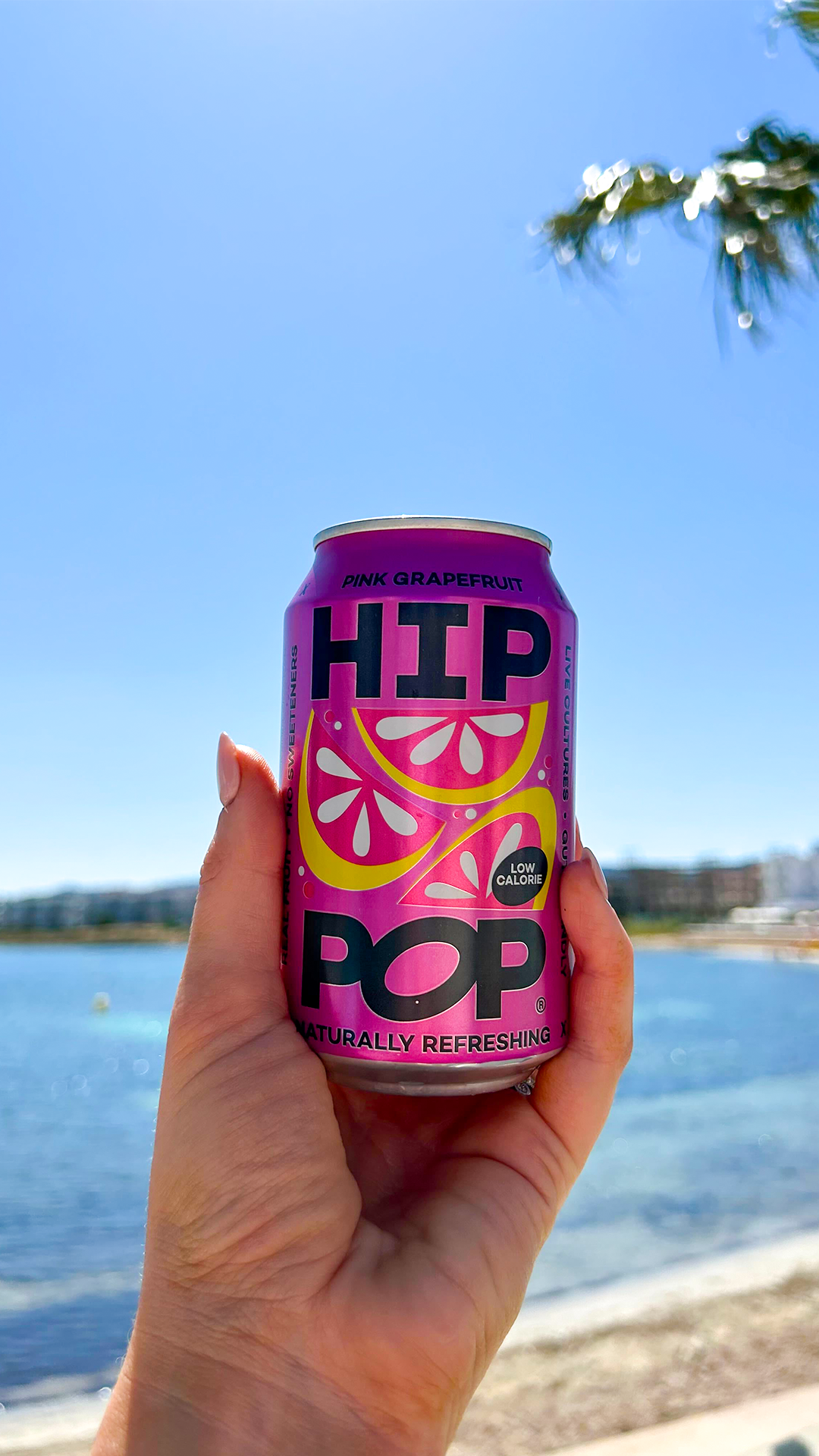 Hand holding a can of pink grapefruit soda by the seaside under a clear blue sky, showcasing a refreshing drink perfect for sunny days.