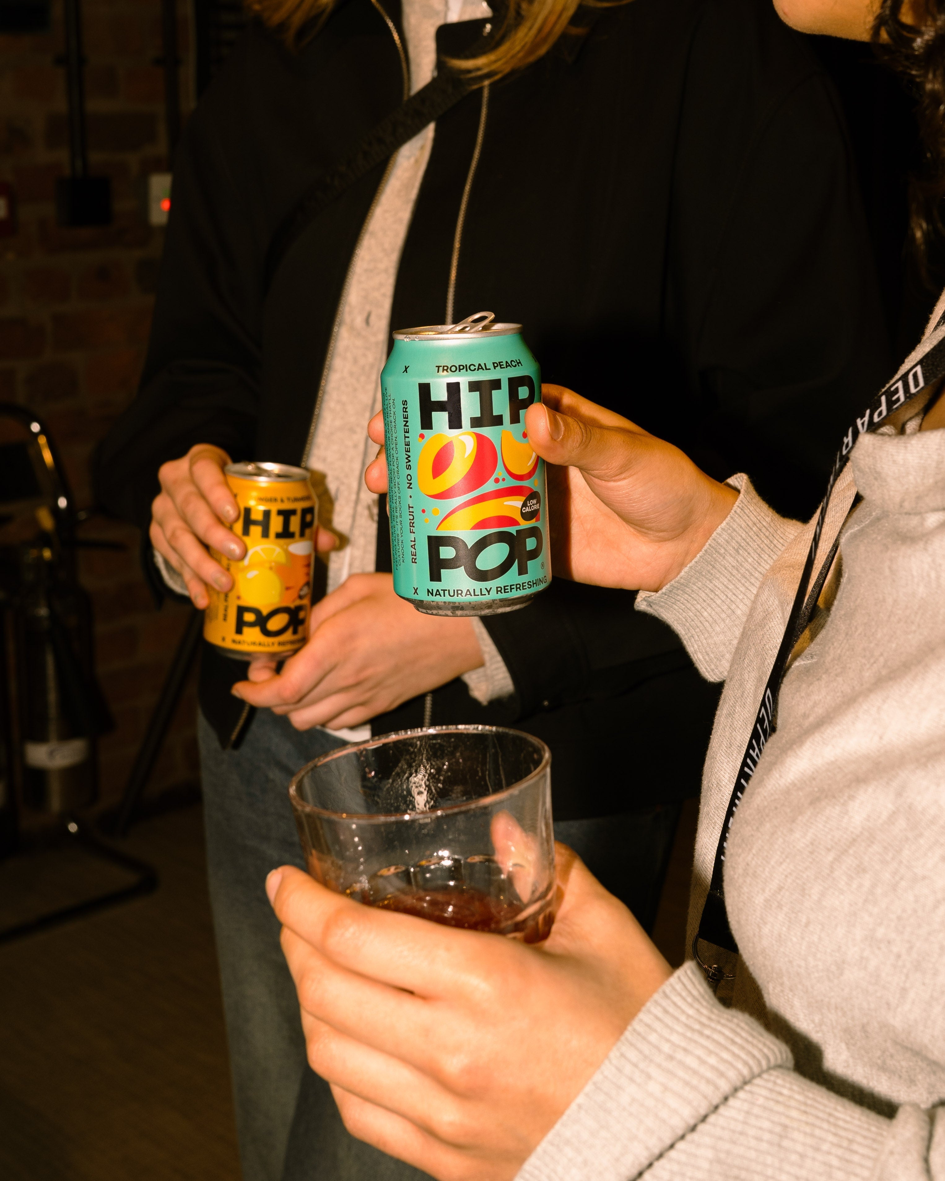 Two people holding cans of Hip Pop; one holding a Tropical Peach flavour can of pop with a vibrant turquoise design, and the other holding a Ginger & Turmeric flavour can with a bright yellow design.