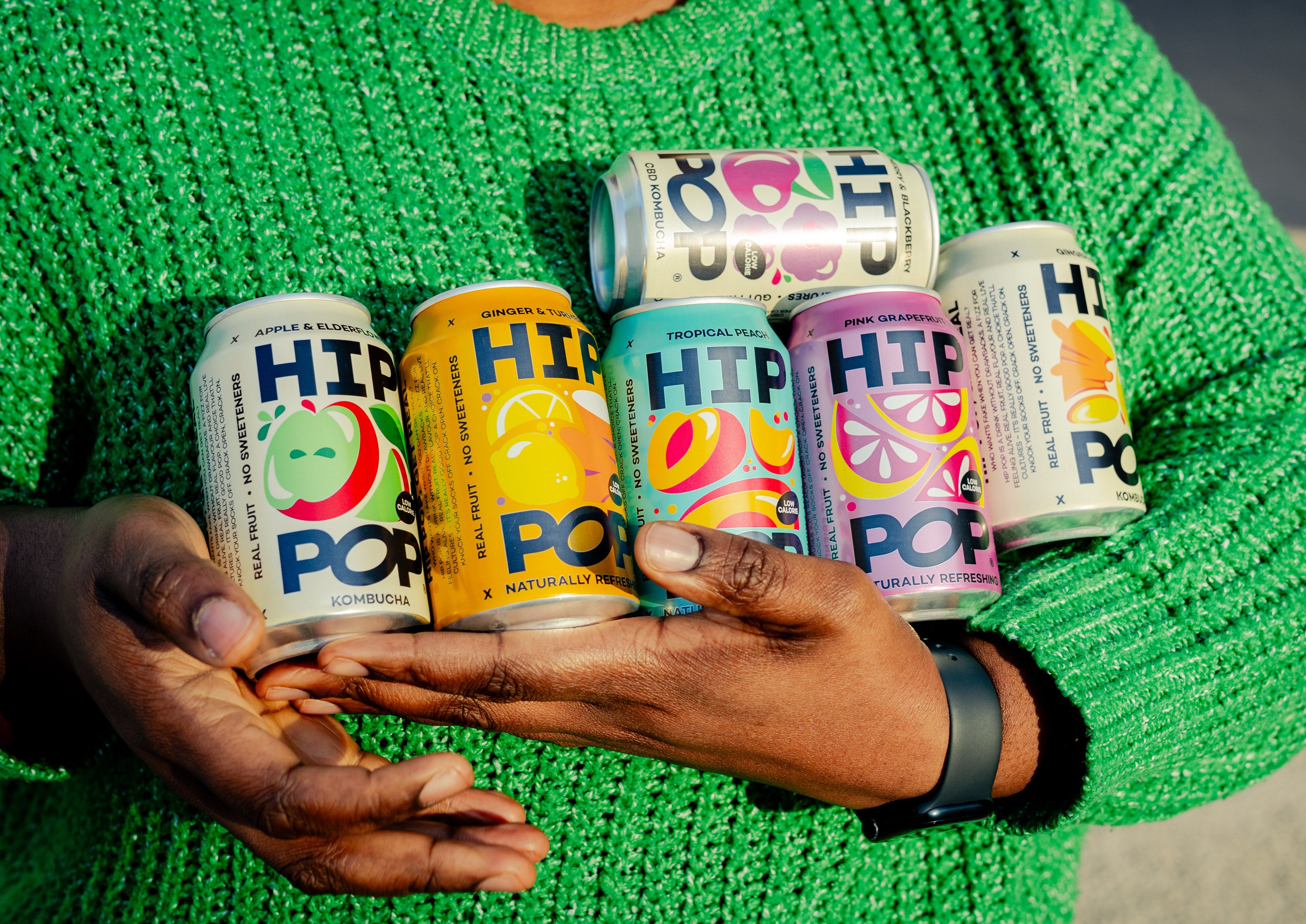 Person in a green jumper holding an assortment of HIP POP kombucha cans, showcasing a colourful range of low calorie drinks made with real fruit and no sweeteners.