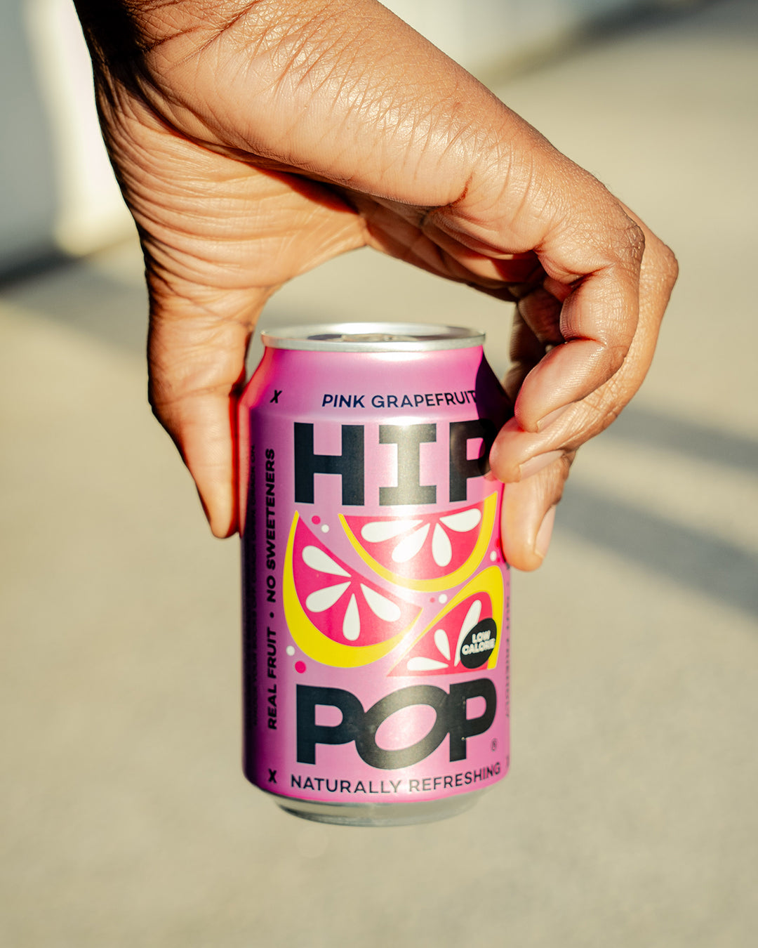 A can of Hip Pop Pink Grapefruit Soda being held, featuring a bold, vibrant design with stylised grapefruit slices and text promoting its natural ingredients and no sweeteners.