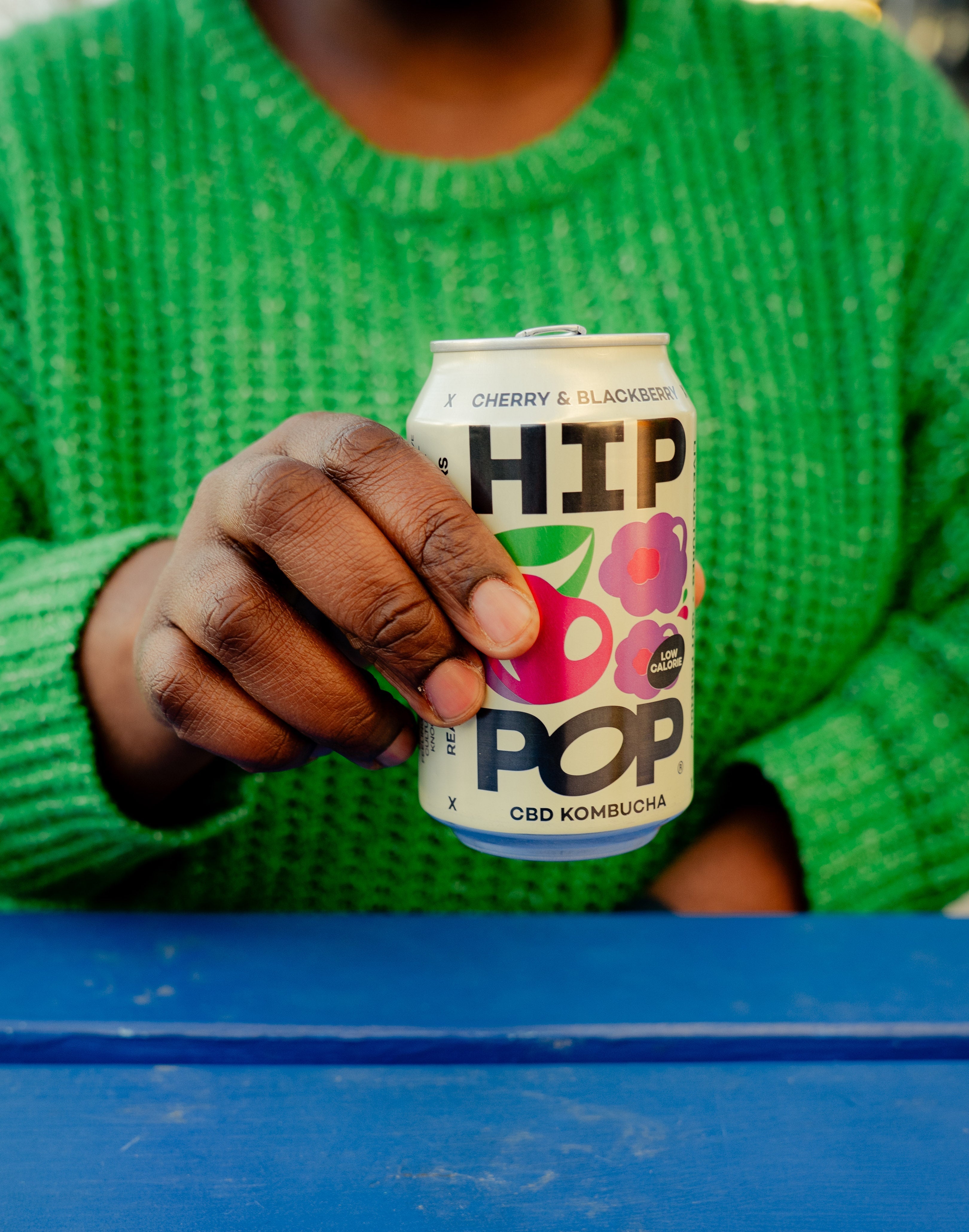 Close-up of a person in a green jumper holding a can of HIP POP cherry and blackberry CBD kombucha, a trendy option among CBD fizzy drinks.
