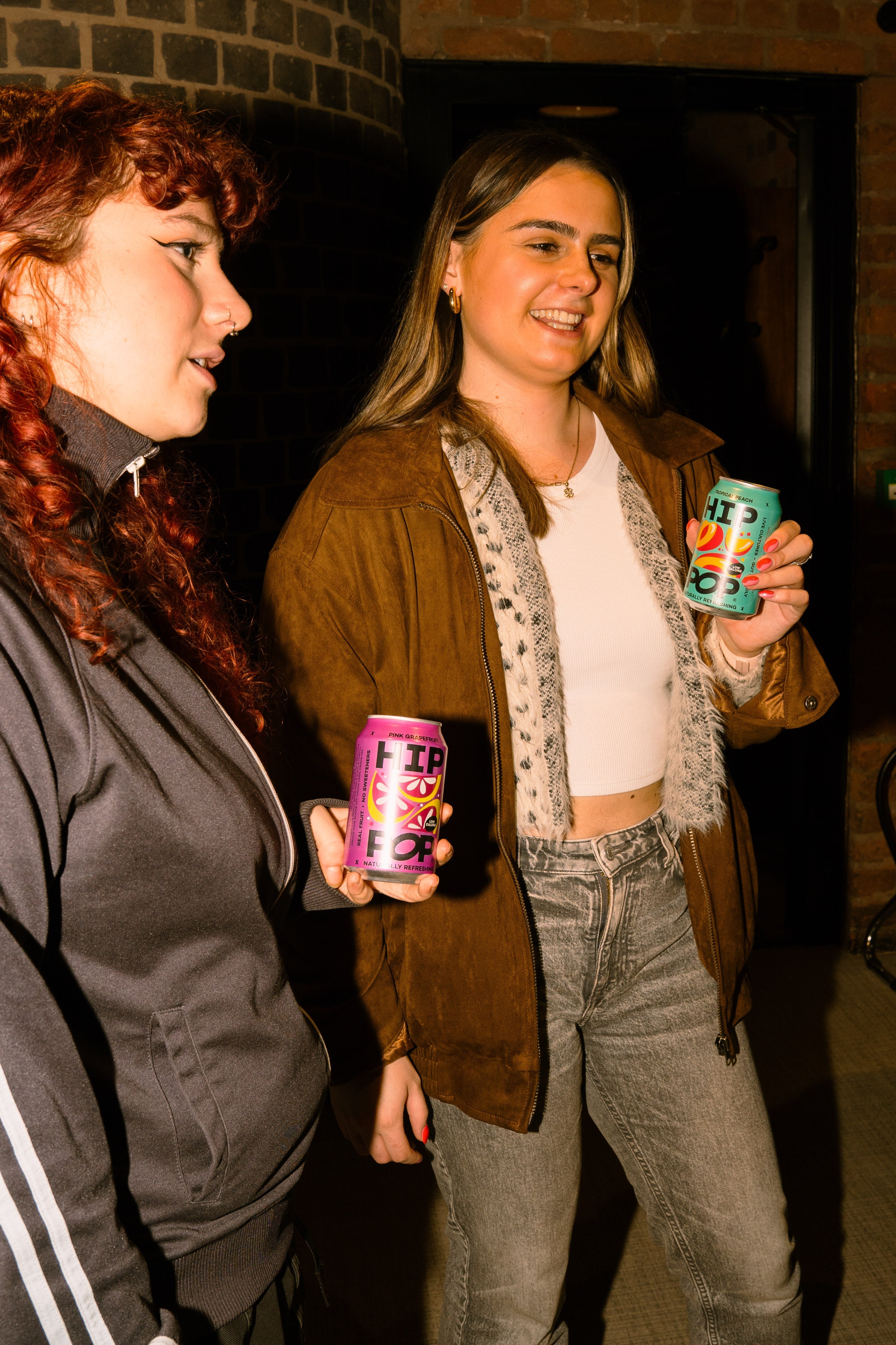 Two young women smiling and holding cans of HIP POP, a drink known for supporting gut health for women with added fibre, while chatting in a warmly lit indoor setting.