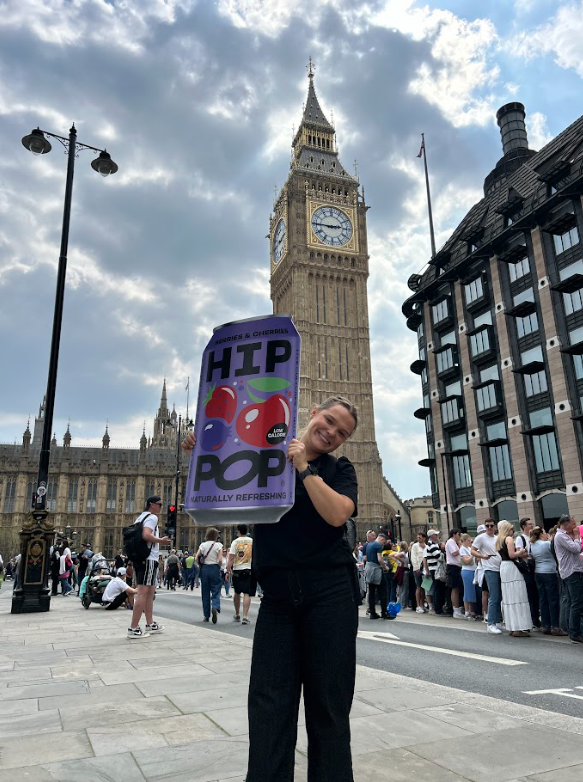 Smiling person holding a giant HIP POP can cut-out in front of Big Ben, promoting gut-friendly drinks as part of discovering how to feel better in my body.