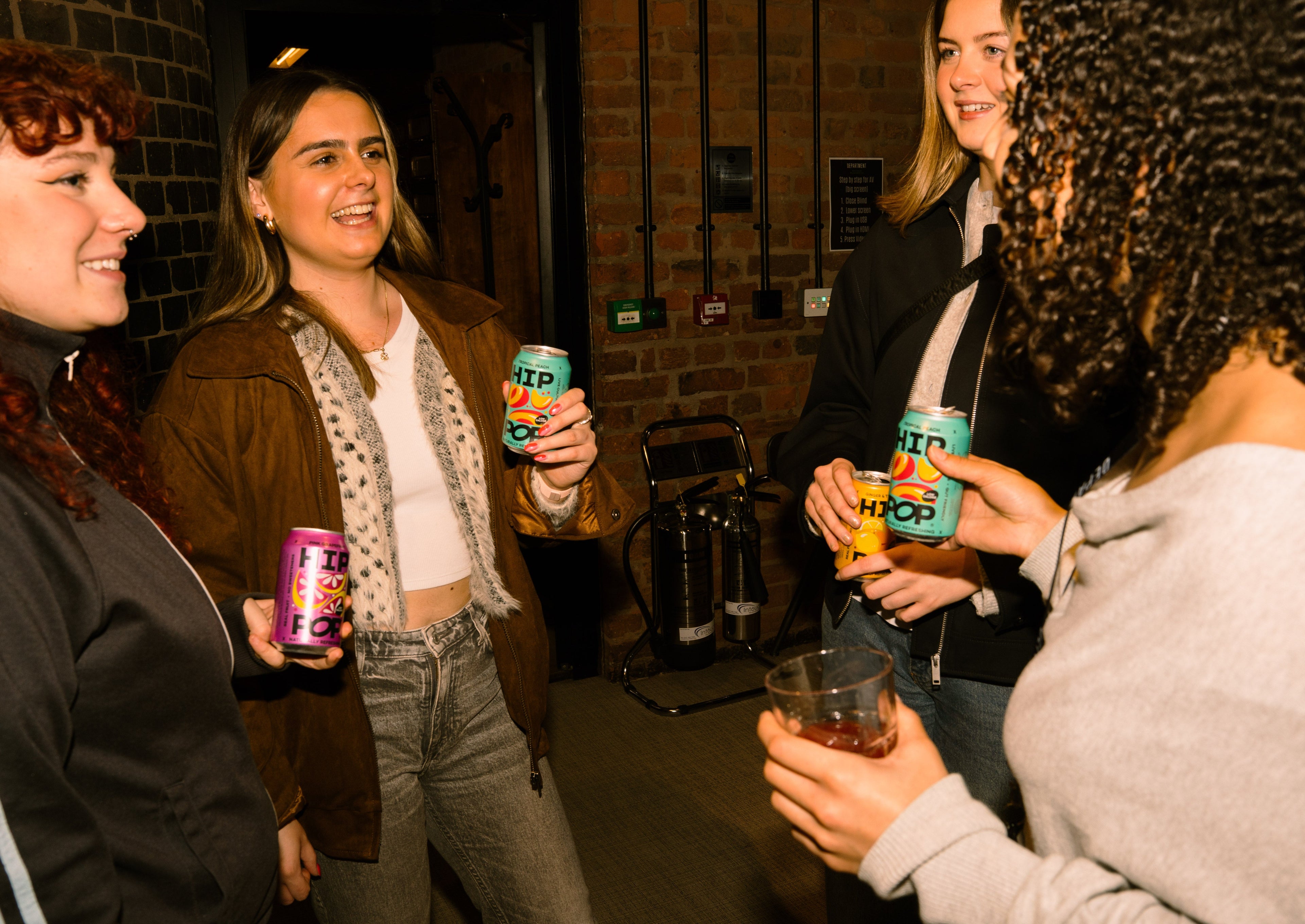 Group of young women enjoying a night out with colourful cans of Hip Pop soda, embracing a sober summer lifestyle in a relaxed indoor setting.