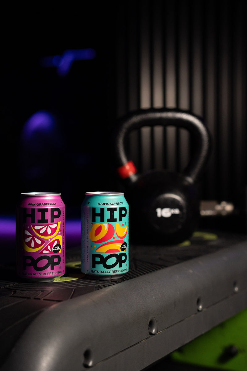 Two vibrant cans of Hip Pop, Pink Grapefruit and Tropical Peach flavours, placed on gym equipment next to a kettlebell, symbolising starting new year’s resolutions early with healthier drink choices.