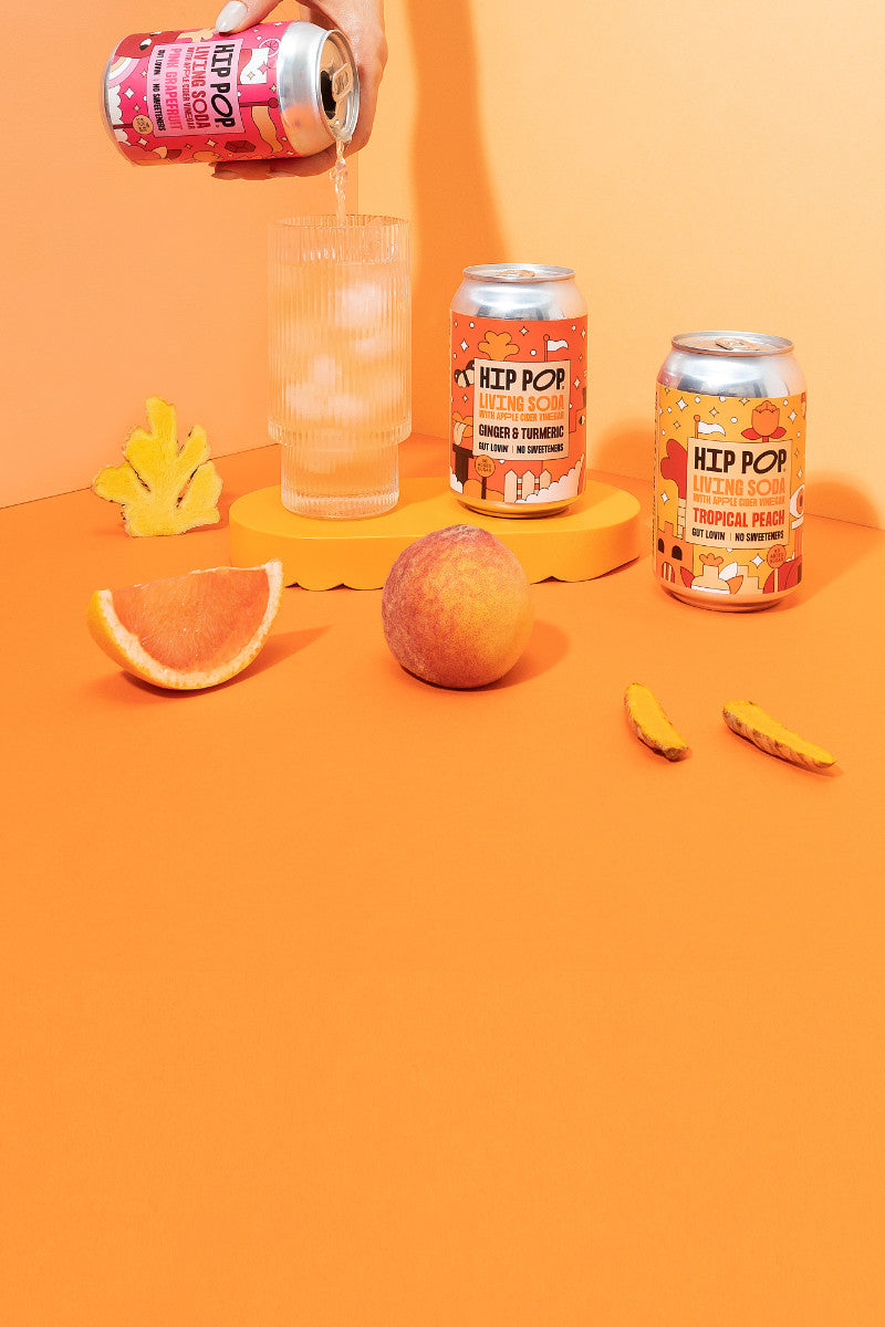 Hip Pop Living Soda collection with fruit and a hand picking up one of the cans and pour into a glass