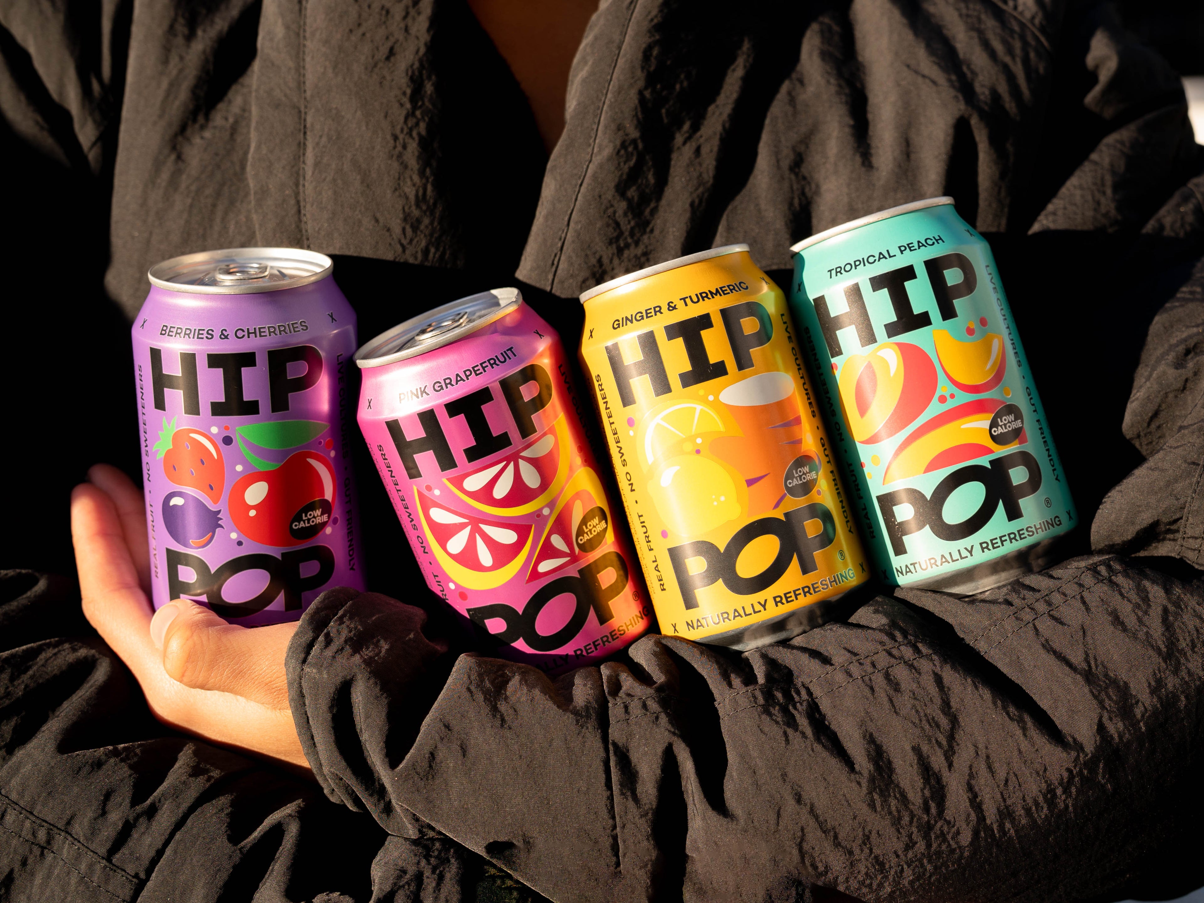 Four colourful cans of HIP POP sweetener-free drinks in assorted flavours, including berries & cherries, pink grapefruit, ginger & turmeric, and tropical peach, held in crossed arms against a dark jacket.