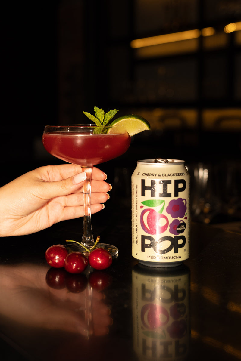 A cocktail glass filled with Hip Pop cherry and blackberry CBD kombucha, a stylish example of functional drinks, garnished with lime and mint beside the matching can and fresh cherries on a bar counter.