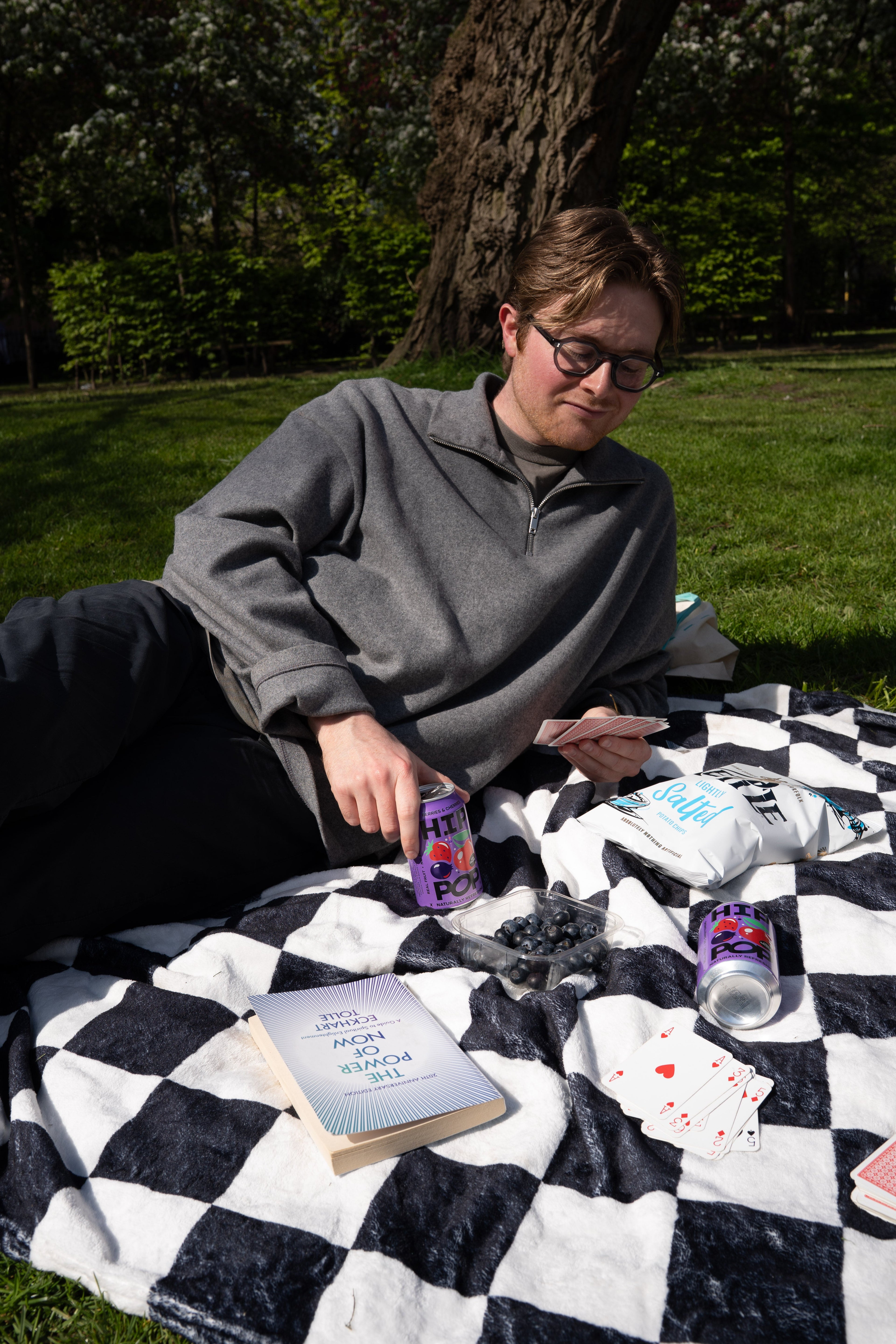 Man relaxing on a picnic blanket with a can of Hip Pop soda and playing cards, enjoying a sunny day in the park; promoting lifestyle habits that support gut health for men.