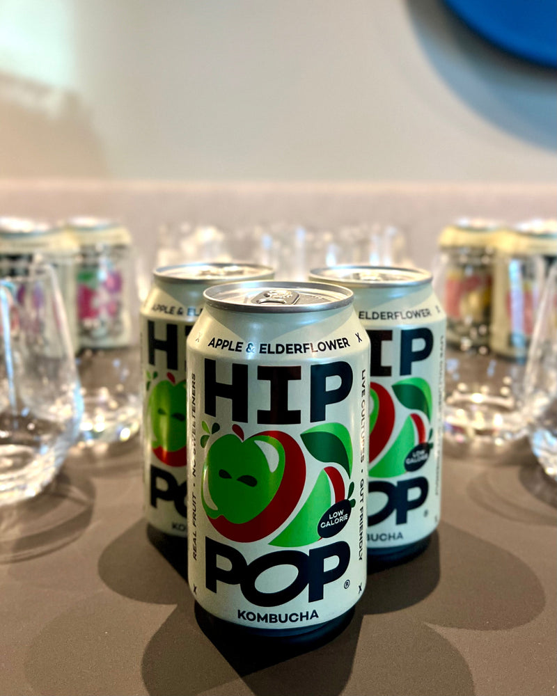 Cans of Hip Pop’s low calorie drinks in apple and elderflower flavour. These three cans of kombucha are displayed on a table with glasses in the background.
