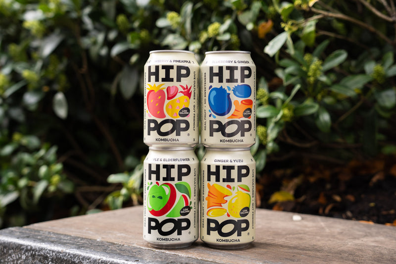 Four cans of HIP POP kombucha stacked outdoors on a wooden surface, featuring colourful fruit illustrations, perfect for celebrating Kombucha Day.