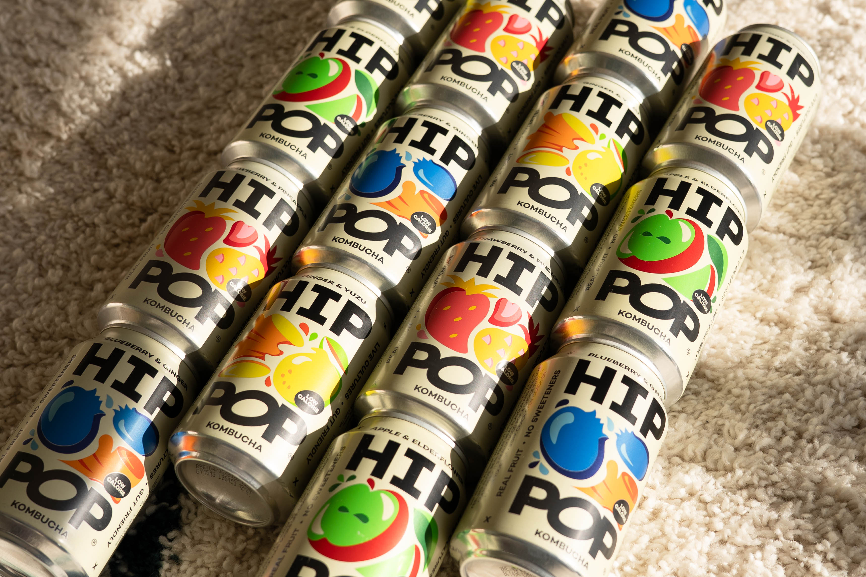 An assortment of Hip Pop kombucha cans featuring vibrant fruit illustrations, arranged diagonally on a soft, textured surface.