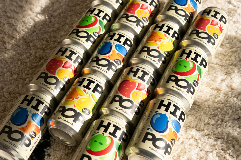 An assortment of Hip Pop kombucha cans featuring vibrant fruit illustrations, arranged diagonally on a soft, textured surface.
