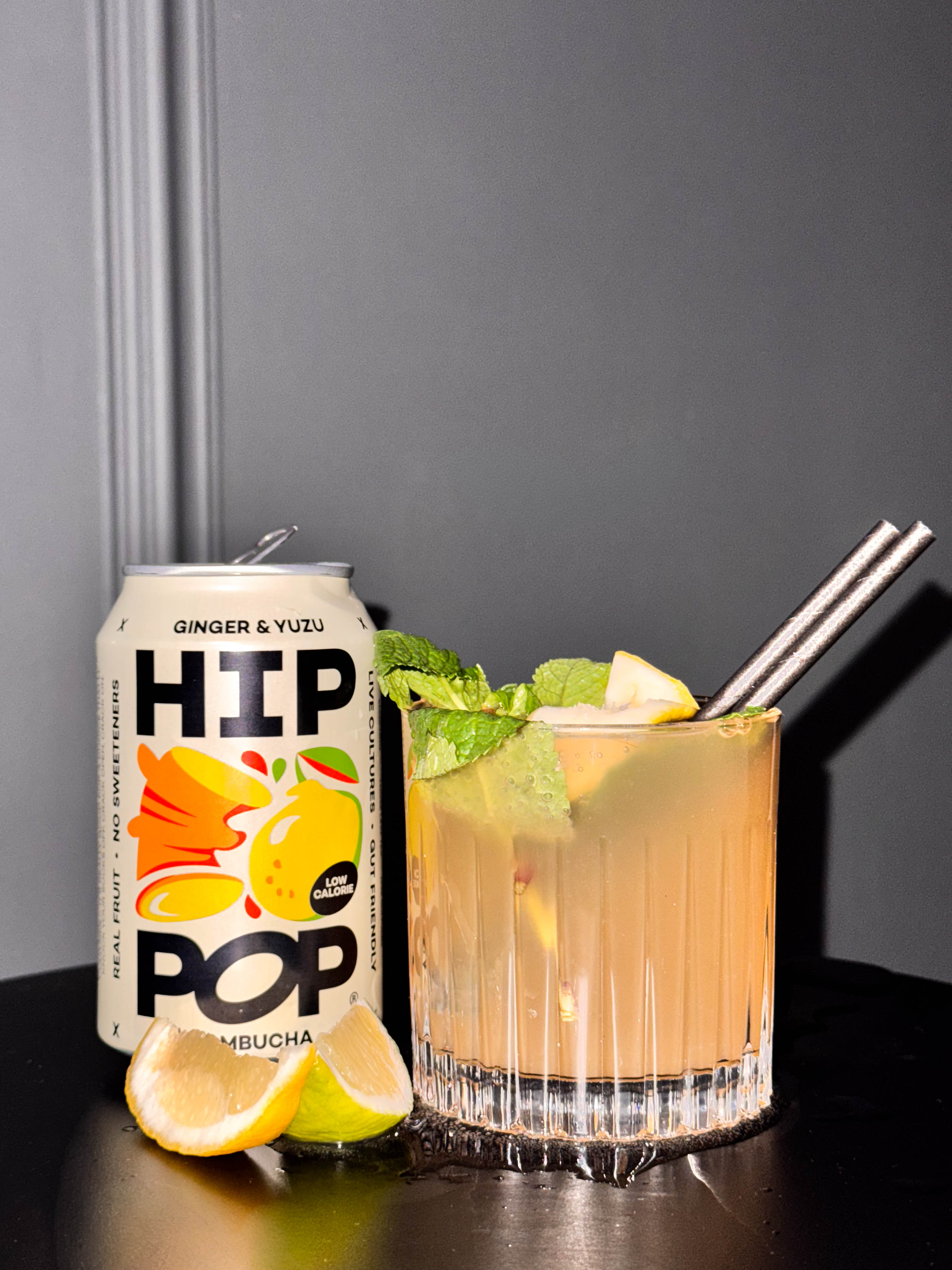  A can of HIP POP Ginger & Yuzu kombucha beside a glass of the chilled drink over ice, garnished with fresh mint and citrus slices; a vibrant choice for sober curious drinks.