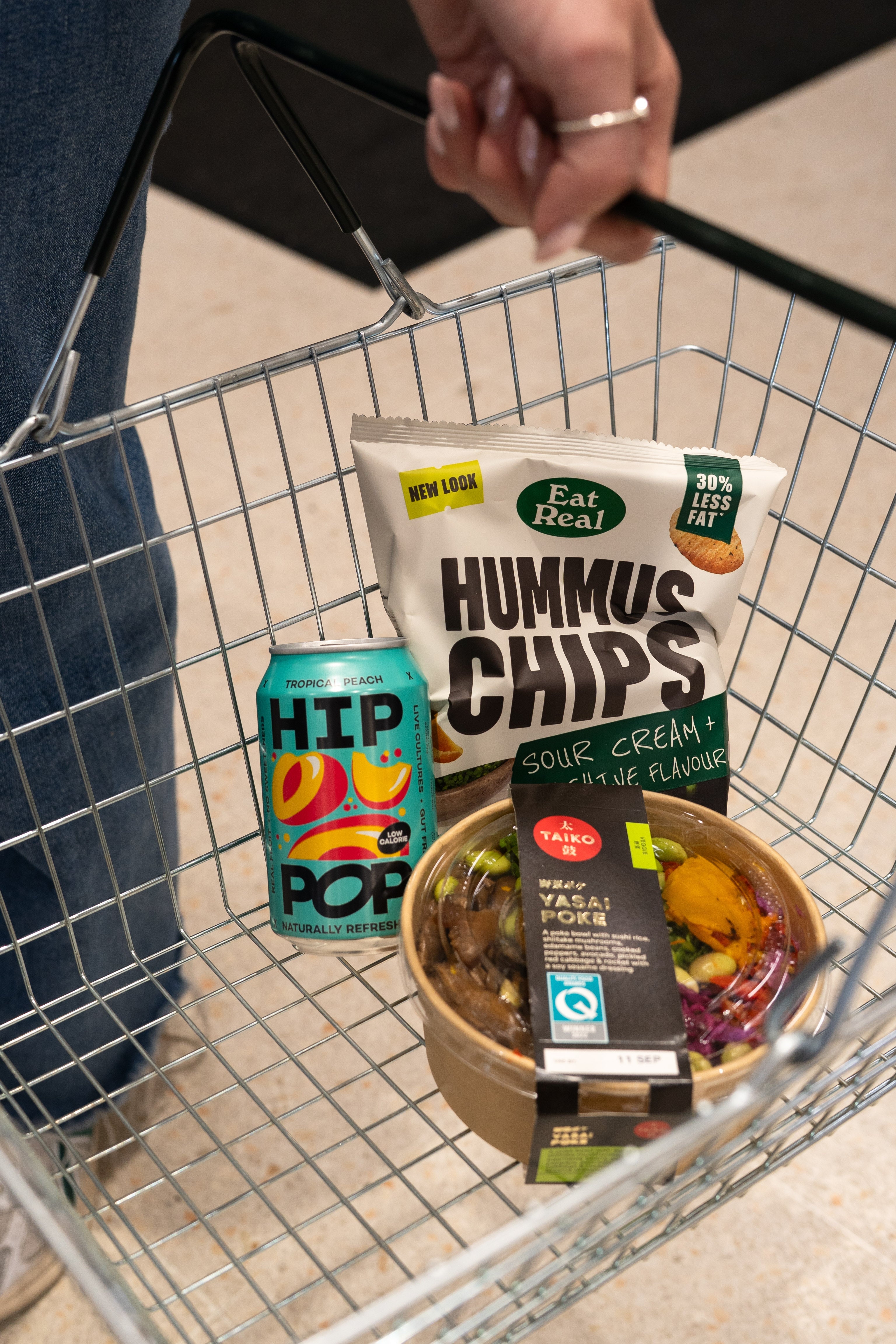 A person holding a shopping basket filled with self-care for winter essentials, including a can of HIP POP kombucha, a bag of sour cream and chive hummus chips, and a vibrant YASAI poké bowl.