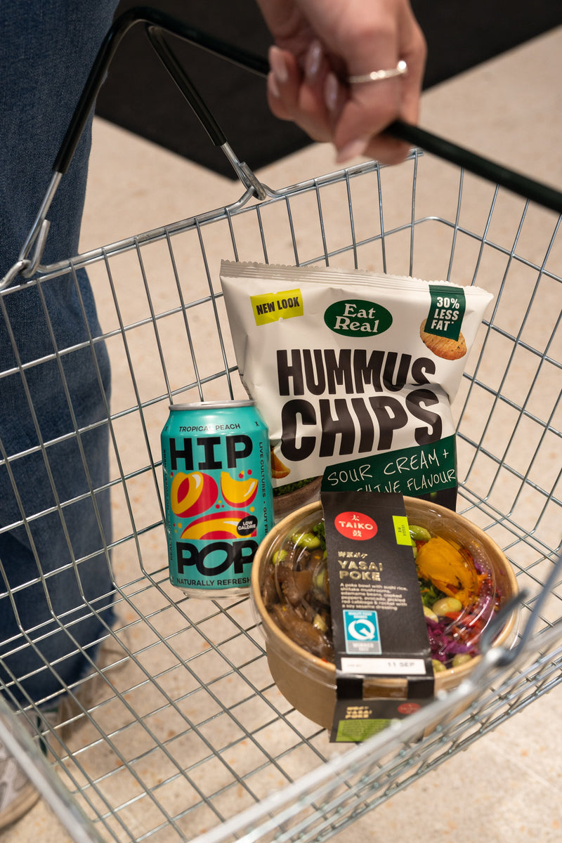 A person holding a shopping basket filled with self-care for winter essentials, including a can of HIP POP kombucha, a bag of sour cream and chive hummus chips, and a vibrant YASAI poké bowl.