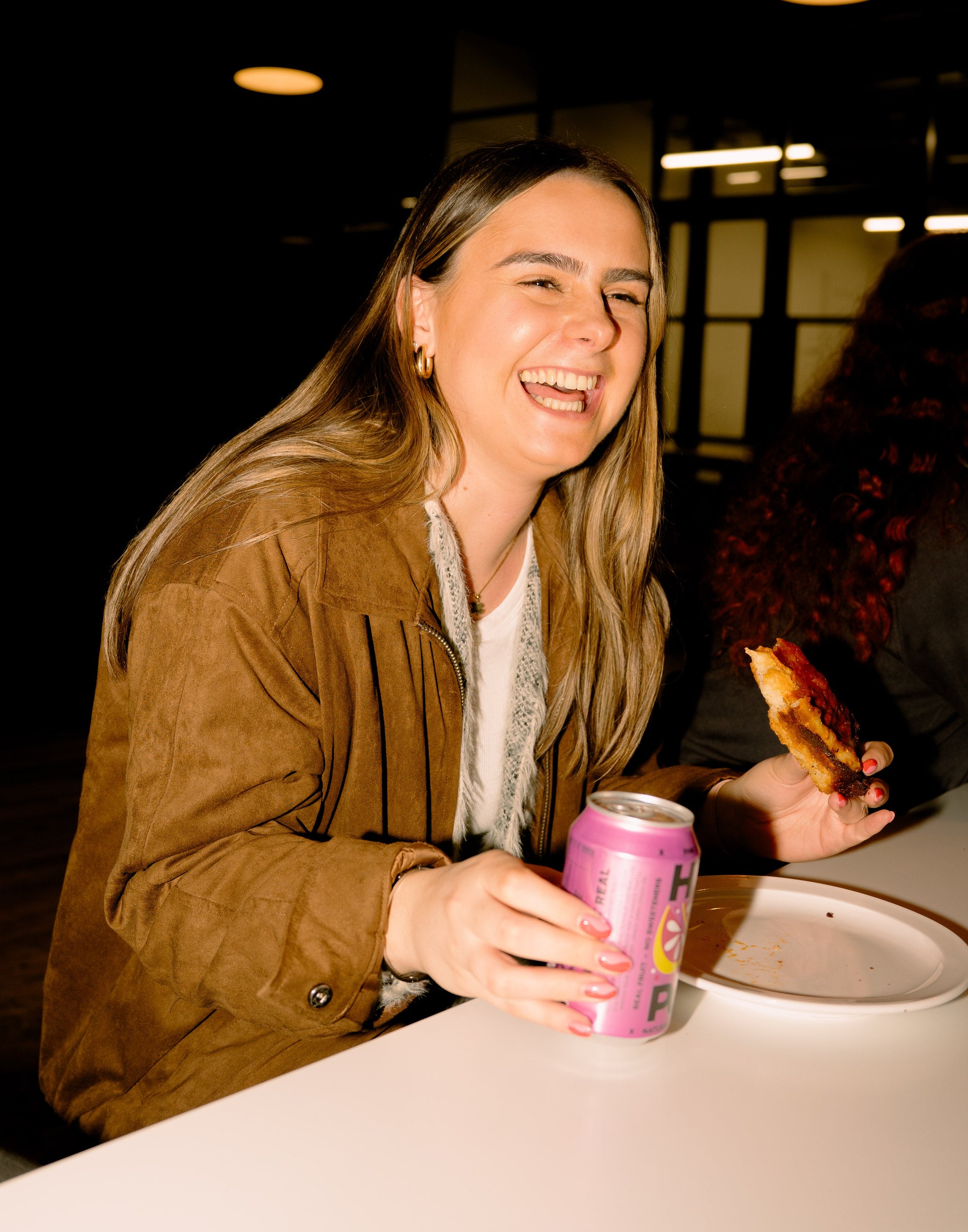 Woman laughing while holding a pink canned drink and a slice of pizza at night, enjoying a relaxed moment despite potential hangxiety.
