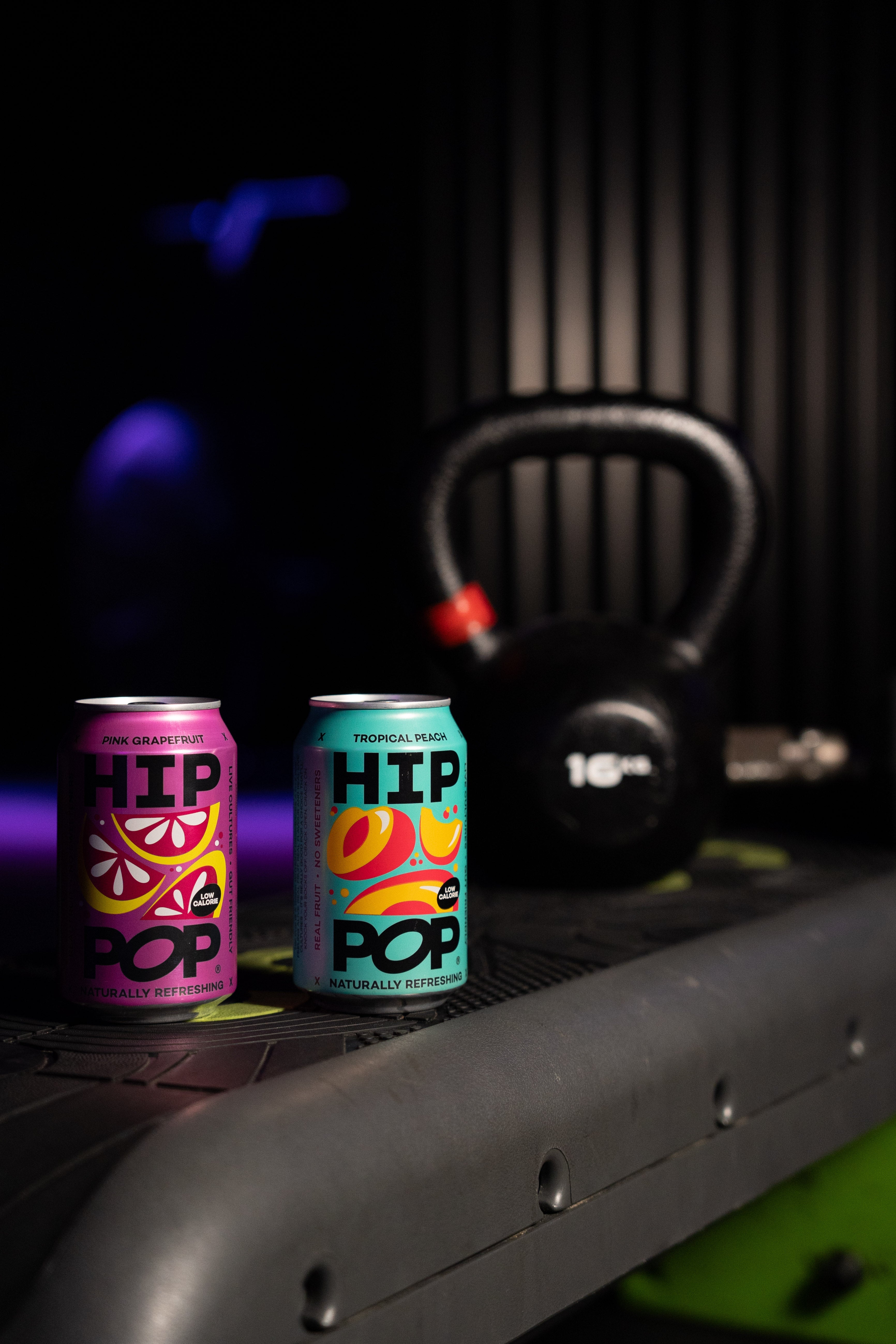 Two vibrant cans of Hip Pop, Pink Grapefruit and Tropical Peach flavours, placed on gym equipment next to a kettlebell, symbolising starting new year’s resolutions early with healthier drink choices.