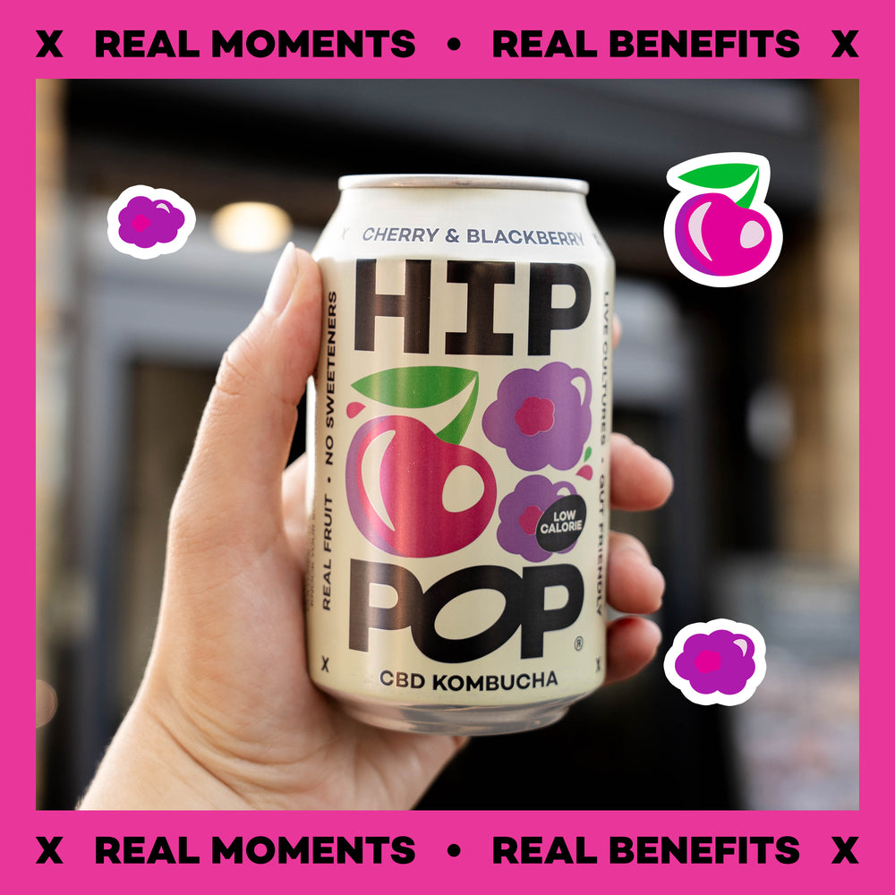 Hand holding a can of HIP POP CBD Kombucha with cherry and blackberry flavor against a pink background.