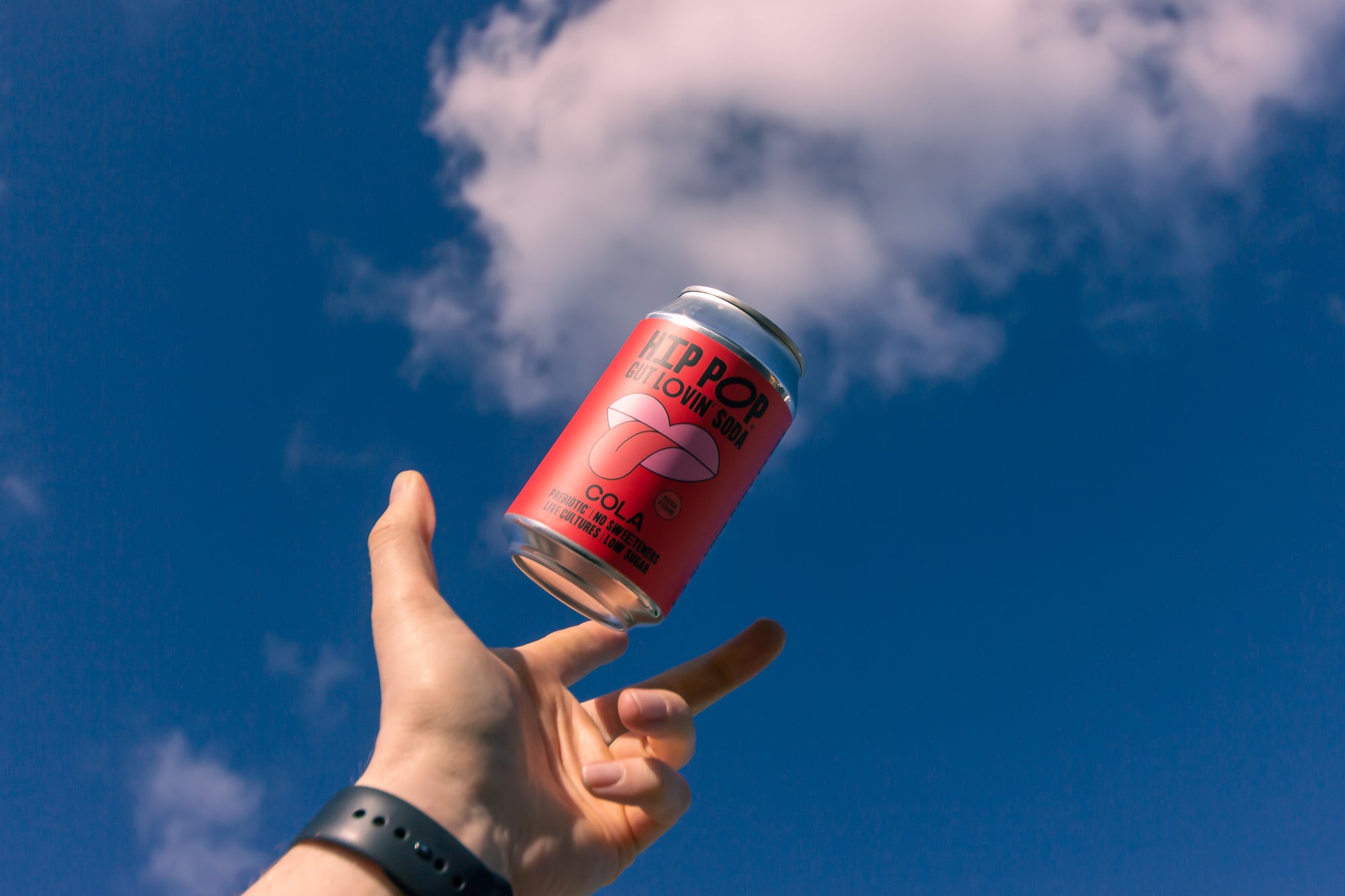 A hand throwing a can of Hip Pop's Gut Lovin' Cola into the air