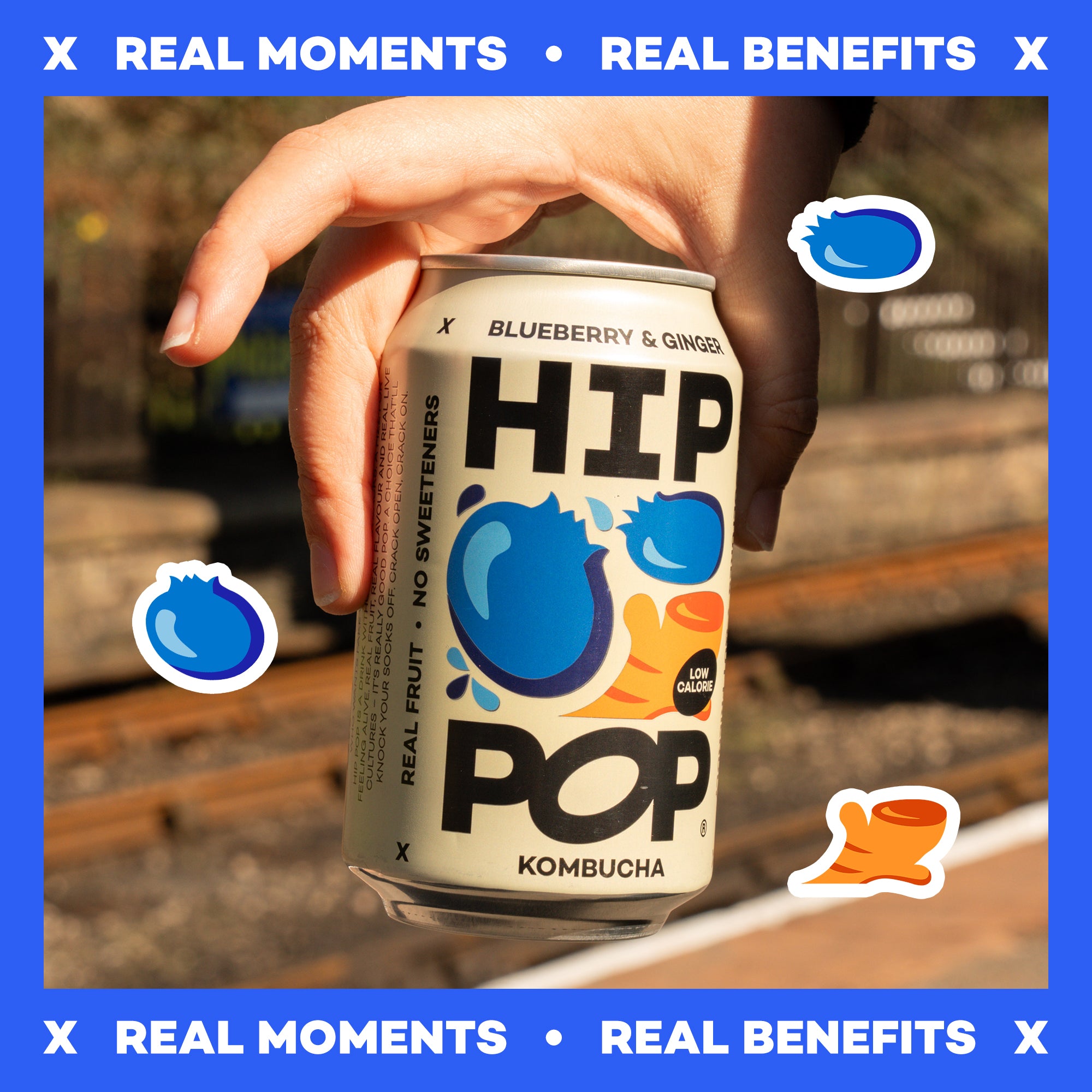 Hand holding a can of HIP Pop Kombucha with blueberry and ginger flavor, set against a blurred outdoor background and text that says "Real Moments" and "Real Benefits".